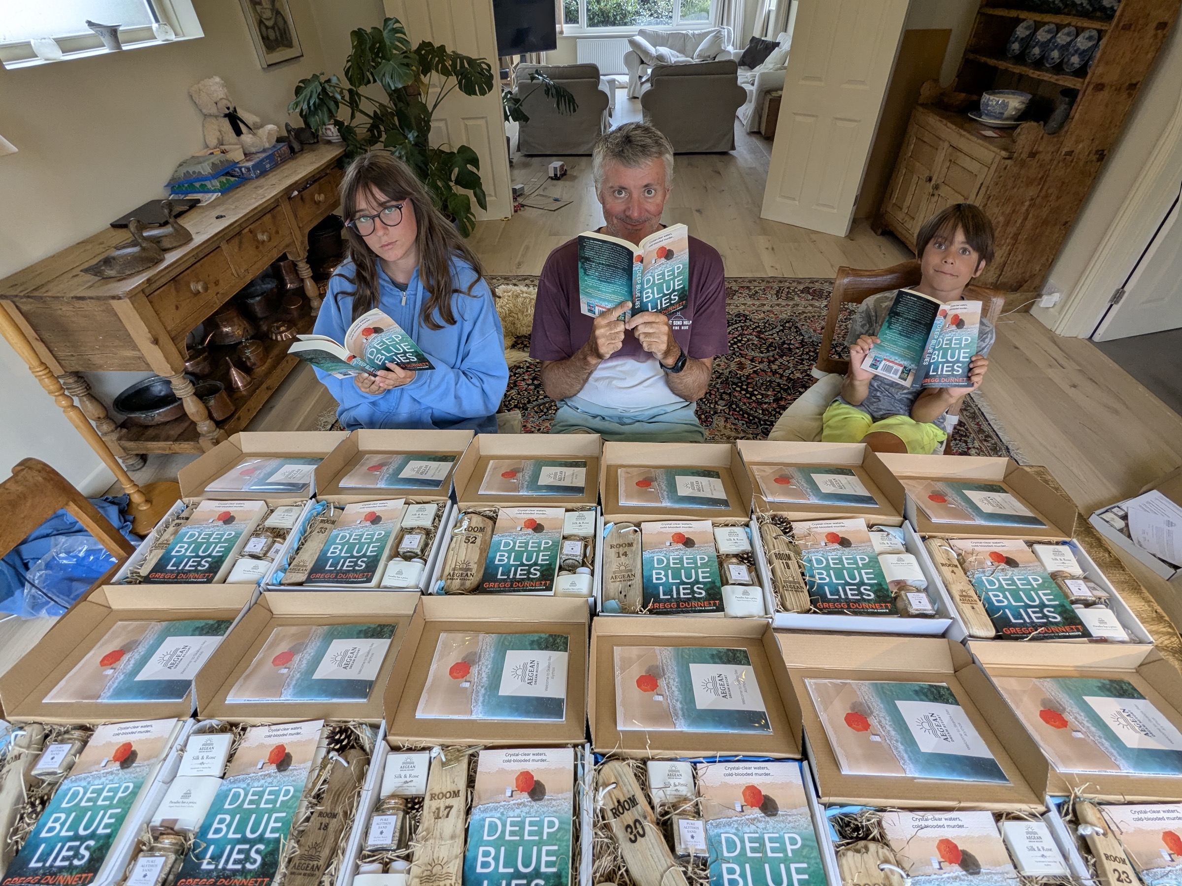 Gregg and his two kids sitting behind a long table, each holding a copy of Deep Blue Lies. The table is laid out with eighteen open collector boxes, each containing the book, an artisan soap, a numbered driftwood room key, and a small jar of sand. Behind them, the family living room.