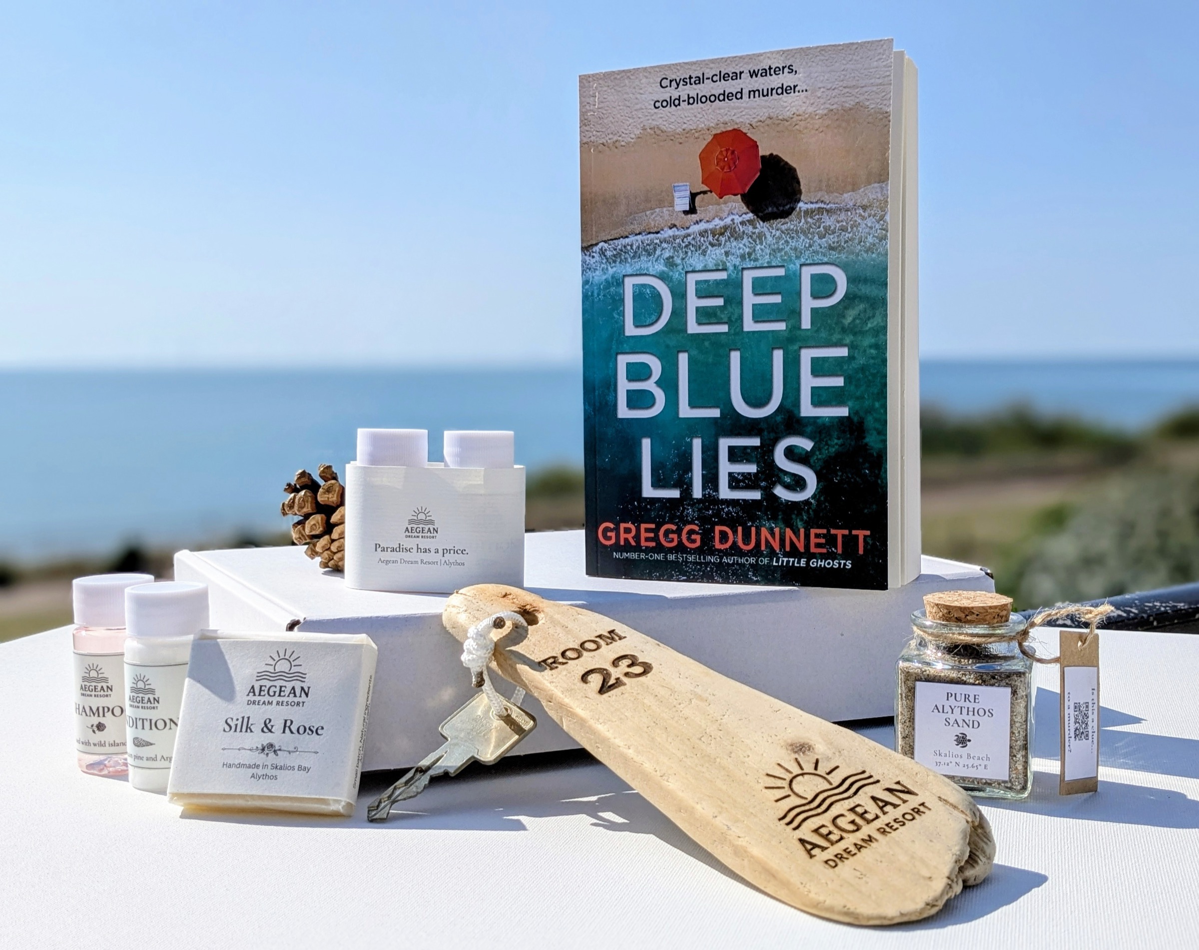 The contents of an Aegean Dream Resort collector box arranged on a white surface against the Cantabrian sea — a numbered driftwood room key labelled 'Room 23', a glass jar of Alythos sand, hand-made soap, miniature shampoo and conditioner bottles, and a paperback copy of Deep Blue Lies.