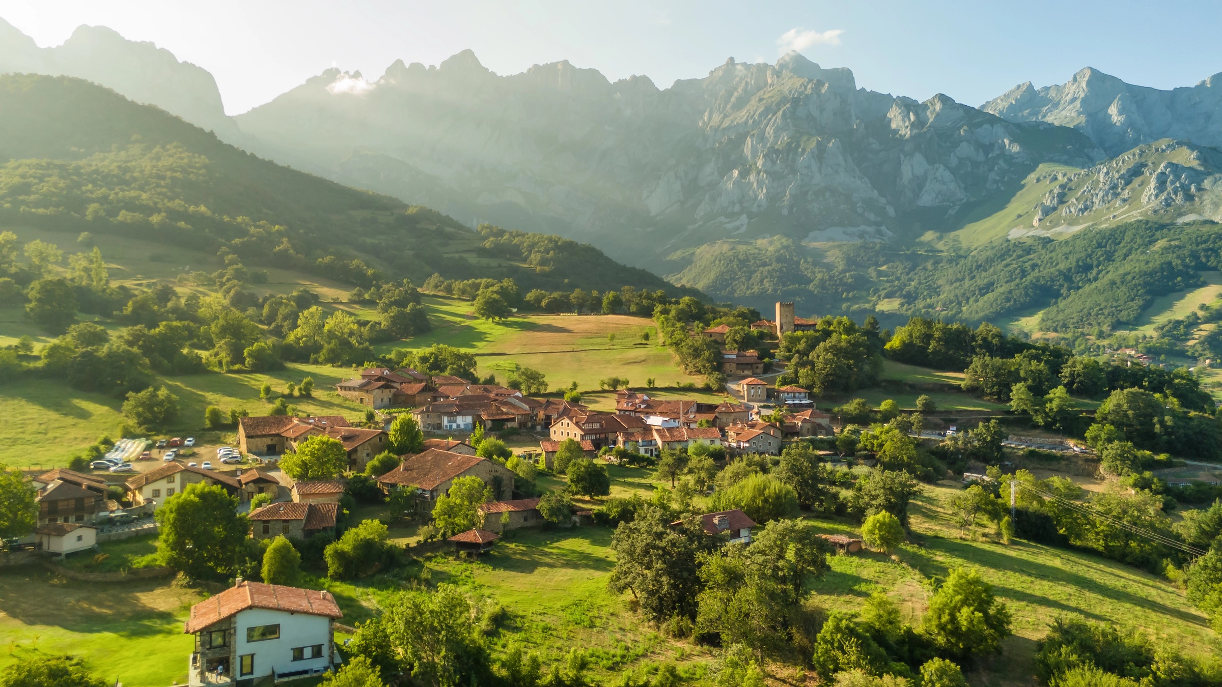 A small Cantabrian village in green pastoral hillsides, with the snow-capped peaks of the Picos de Europa rising behind it in low evening sun.